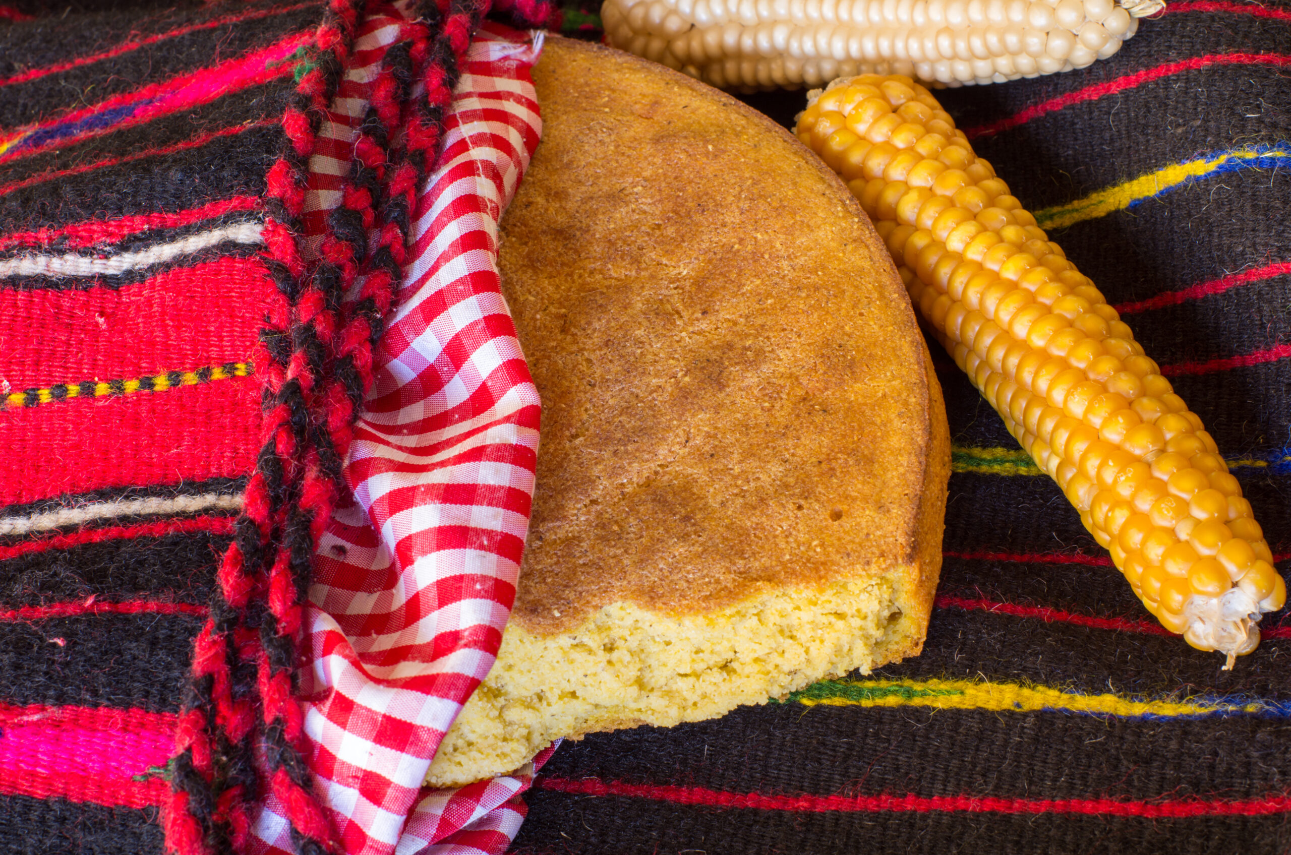 Traditional Montenegrin cornbread (kukuruza) partially wrapped in a red-checked cloth and colorful woven fabric, with yellow and white corn cobs beside it.
